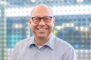 Ein lächelnder, kahlköpfiger Mann mit Brille und blau gestreiftem Hemd vor einem verschwommenen Hintergrund aus geometrischen Formen und einem Hauch von Grün. Sein fröhlicher Gesichtsausdruck vermittelt Freundlichkeit und Zugänglichkeit.