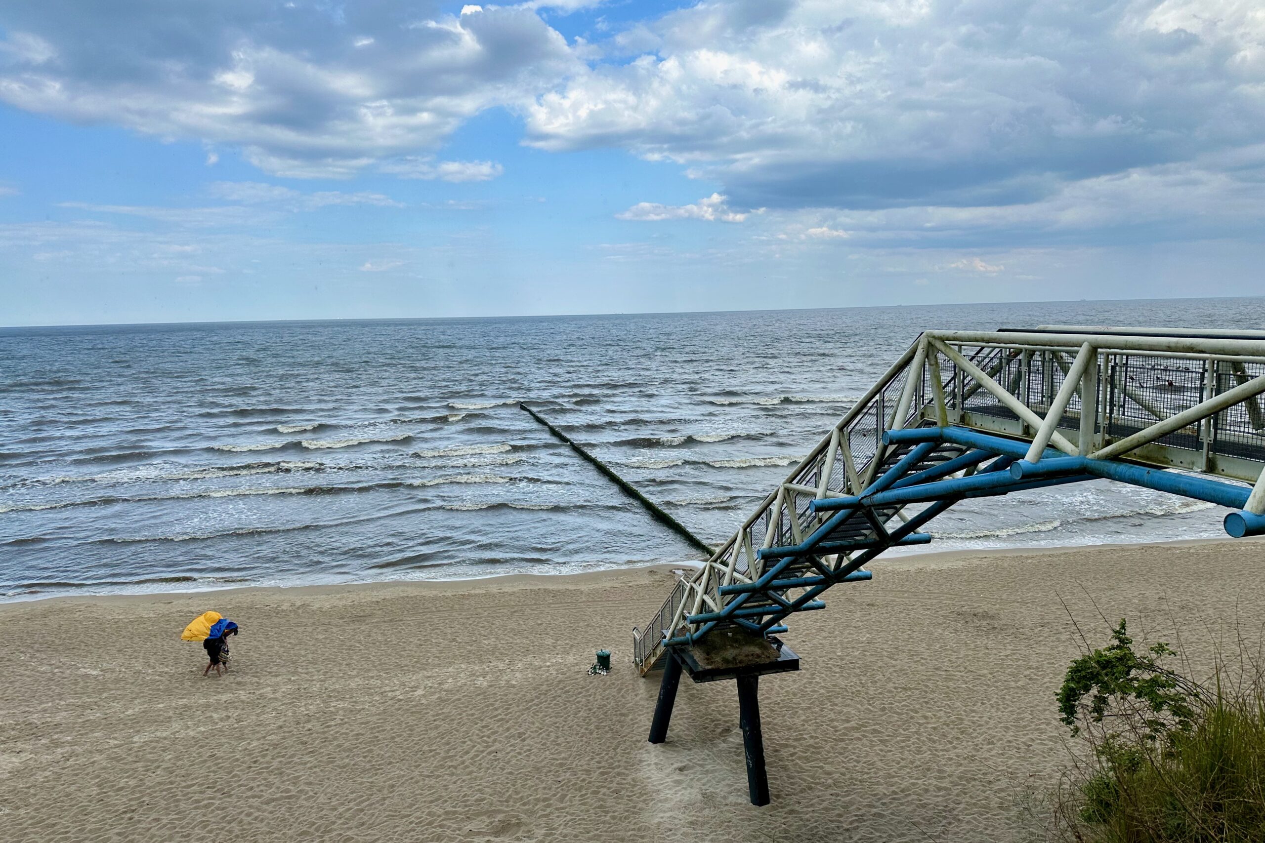 Eine Person mit einem gelben Regenschirm geht an einem Sandstrand am Meer in Usedom entlang. Die Wellen nähern sich sanft dem Ufer und ein langer Pier mit einer Metallkonstruktion ragt teilweise über Wasser und Sand hinaus. Der Himmel ist voller Wolken, die einen grauen Farbton über die Szene werfen. Auf der rechten Seite ist spärliches Grün zu sehen.