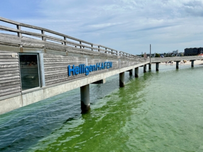 Ein langer Holzsteg erstreckt sich &uuml;ber gr&uuml;nliches Meerwasser unter einem teilweise bew&ouml;lkten Himmel. Auf der Seite des Stegs steht in blauen Buchstaben &bdquo;Heiligenhafen&ldquo;. Im Hintergrund sind ein Sandstrand mit Menschen und Geb&auml;uden zu sehen, was eine K&uuml;stenstadt vermuten l&auml;sst.