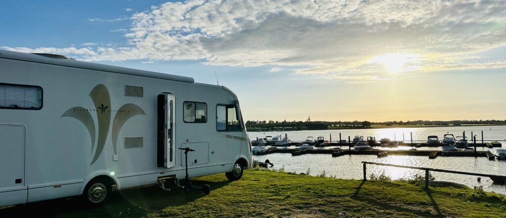 Wohnmobil BLUΞFIRΞ ON TOUR, steht auf einer Rasenfläche in Ufernähe. Die untergehende Sonne wirft warmes Licht auf die Szene, wo ein paar Wolken den blauen Himmel sprenkeln und Boote am Ufer angedockt sind.