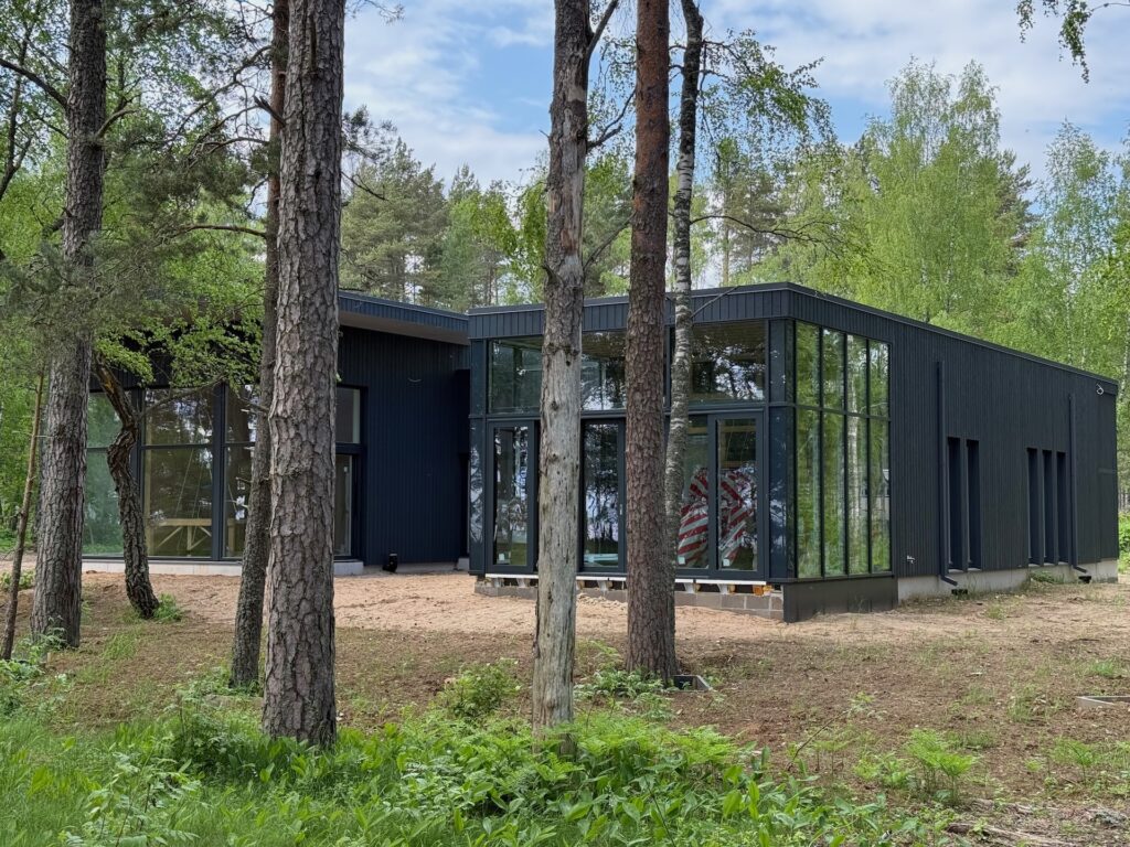 Baltikum 2025 – Lettland 2 – 13 Ein modernes schwarzes Haus mit großen Glasfenstern steht zwischen hohen Bäumen und viel Grün und fügt sich unter einem teilweise bewölkten Himmel in die natürliche Waldumgebung ein.