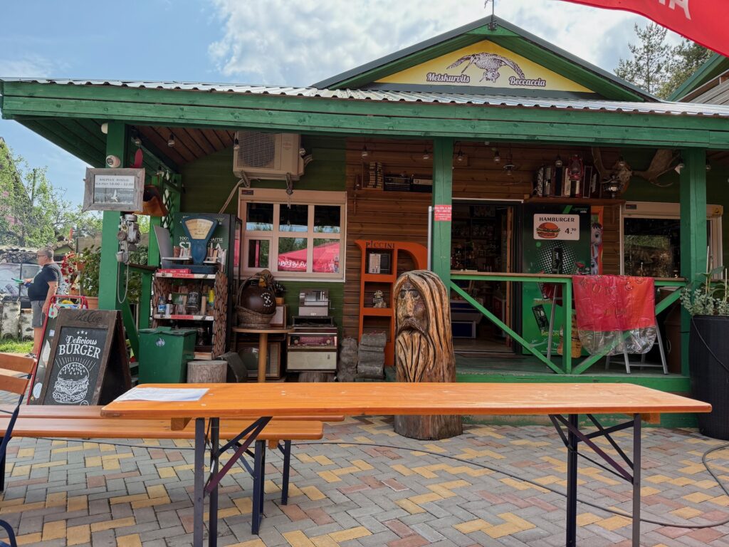 Baltikum 2025 – Lettland 2 – 14 Ein kleines Café aus grünem Holz mit einer überdachten Veranda, Bärenschnitzereien aus Holz, einer Kreidetafel mit Burger-Menü, Picknicktischen im Freien und farbenfroher Dekoration in einem sonnigen Innenhof mit Ziegelpflaster.