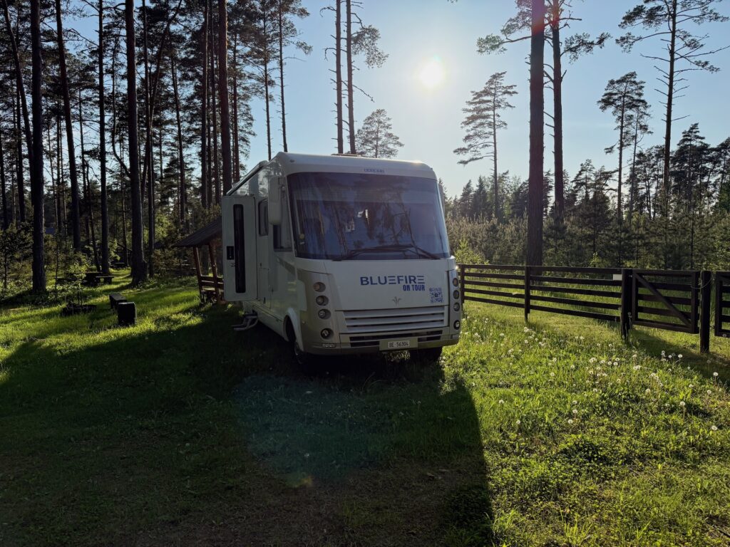 Baltikum 2025 – Lettland 2 – 15 Ein weißer Wohnwagen mit der Aufschrift „BLUEFIRE“ steht auf einer Wiese auf einer sonnenbeschienenen Waldlichtung, umgeben von hohen Bäumen und einem Holzzaun, durch dessen Äste Sonnenlicht fällt.