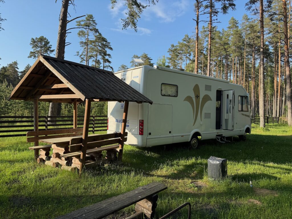 Baltikum 2025 – Lettland 2 – 16 Ein weißer Wohnwagen steht auf einer Wiese in der Nähe eines hölzernen Picknickplatzes und einer Bank in einem Waldgebiet mit hohen Kiefern unter blauem Himmel.