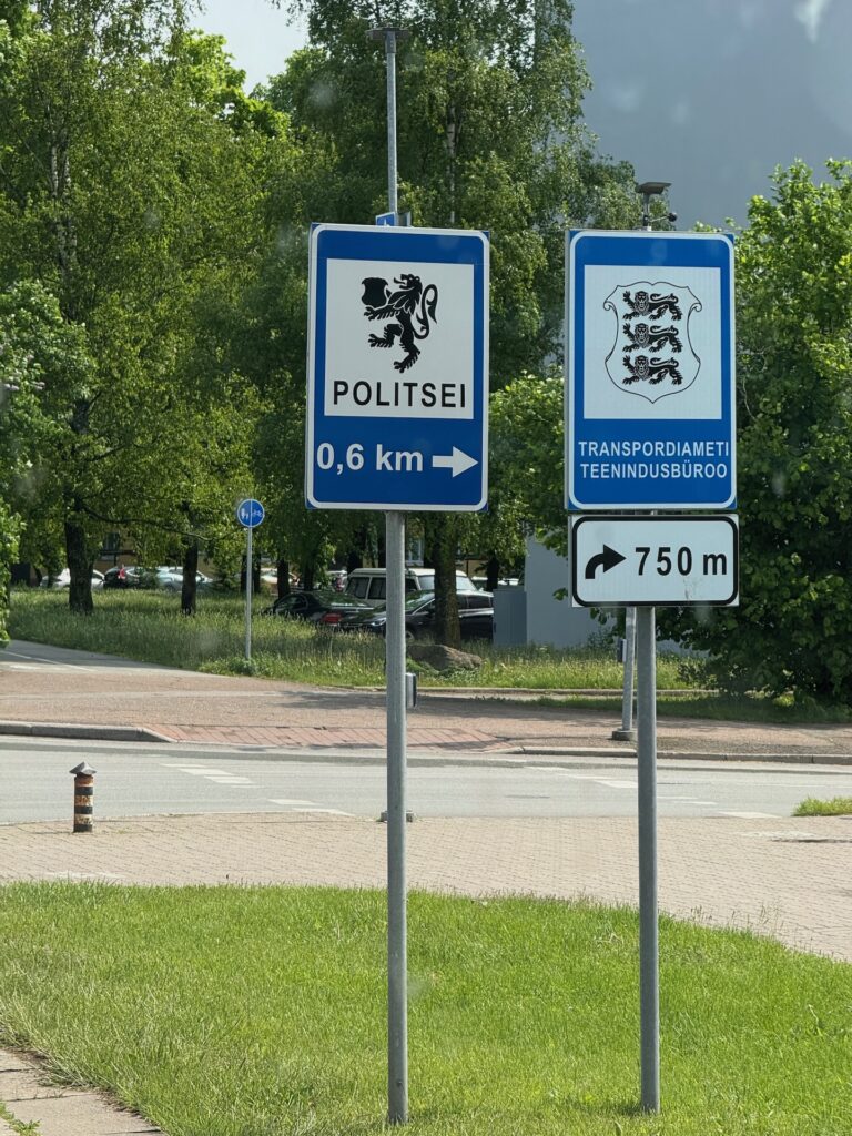 Baltikum 2025 – Lettland 2 – 18 Zwei estnische Verkehrsschilder weisen nach rechts: Eines zeigt an, dass die Polizeistation 0,6 km entfernt ist, das andere, dass das Straßenverwaltungsbüro 750 Meter entfernt ist. Beide Schilder tragen schwarze Löwen als Wappen. Im Hintergrund sind Bäume und Gras zu sehen.