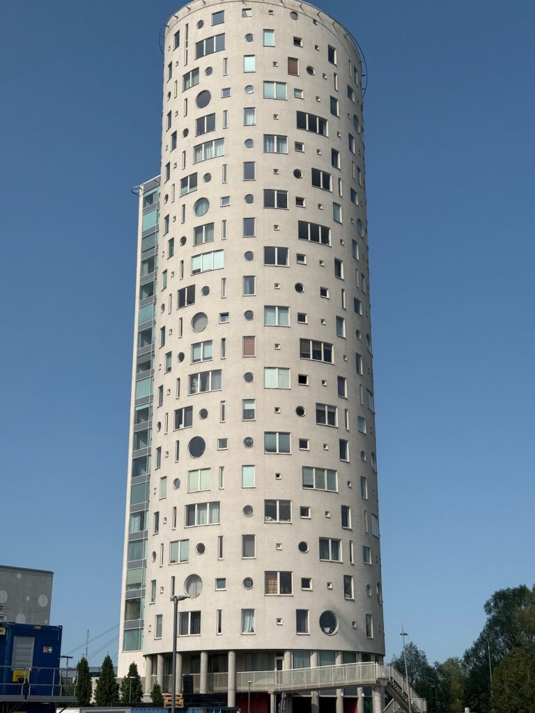Baltikum 2025 – Lettland 2 – 2 Vor einem klaren blauen Himmel erhebt sich ein hohes, zylindrisches Wohnhaus mit zahlreichen Fenstern unterschiedlicher Größe und Form an der hellgrauen Fassade. In der Nähe befinden sich einige Bäume und kleinere Gebäude.