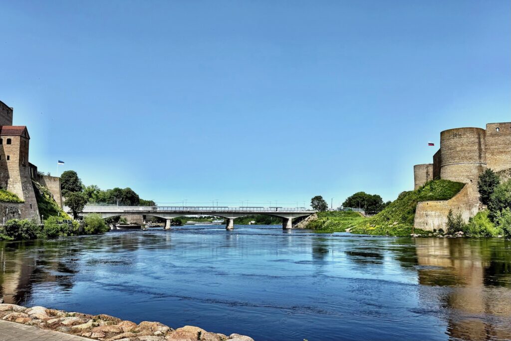 Baltikum 2025 – Lettland 2 – 20 Ein ruhiger Fluss fließt unter einer Betonbrücke hindurch, mit historischen Steinfestungen an beiden Ufern, grünem Gras und Bäumen an einem klaren, sonnigen Tag.