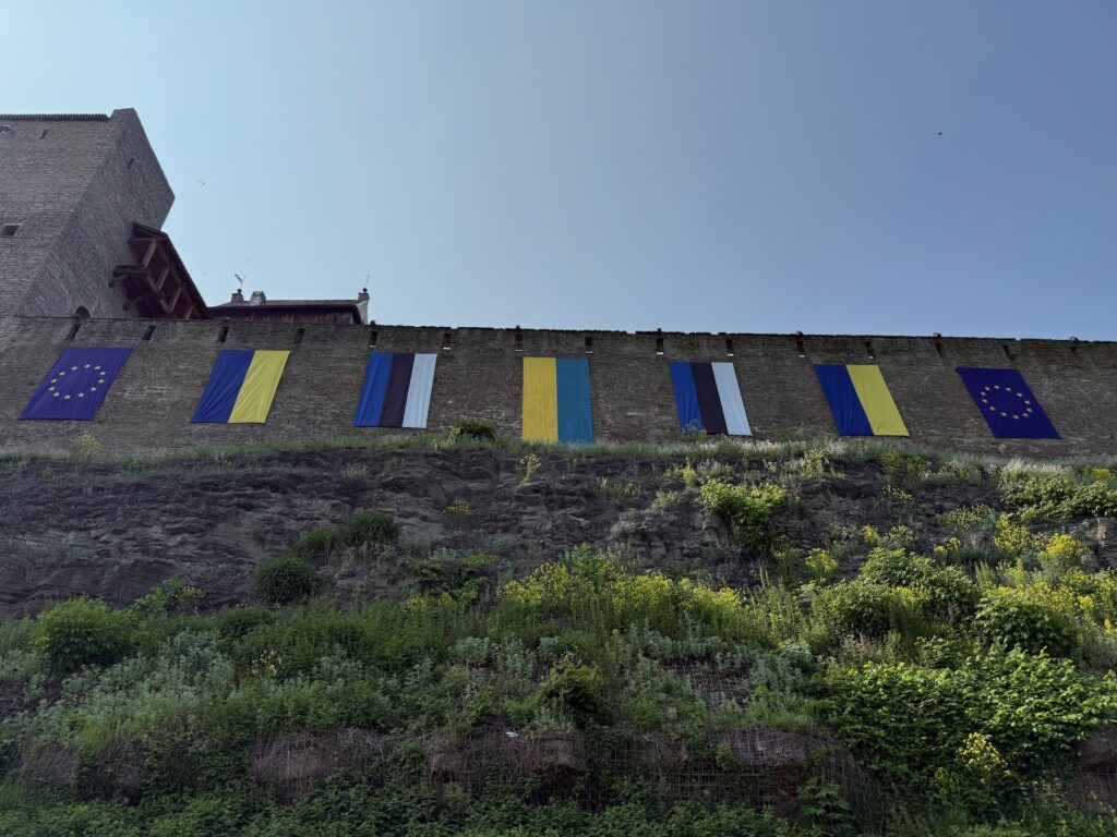 Baltikum 2025 – Lettland 2 – 22 Mehrere Flaggen, darunter die Flagge der Europäischen Union, der Ukraine und Estlands, hängen unter einem klaren blauen Himmel an einer Steinmauer über einem grasbewachsenen Hang.