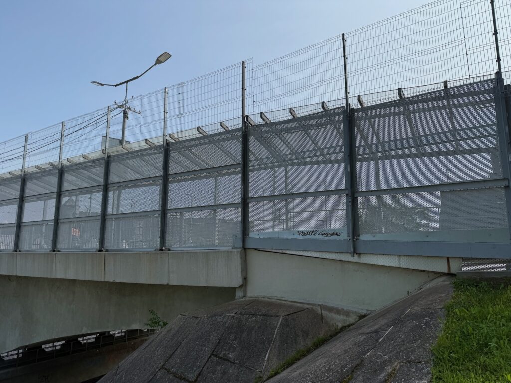 Baltikum 2025 – Lettland 2 – 23 Eine Betonbrücke mit einem hohen Metallzaun und einem Netz darüber, vermutlich aus Sicherheitsgründen, überspannt einen kleinen Kanal. Vor einem klaren blauen Himmel sind Straßenlaternen und Stromleitungen zu sehen.