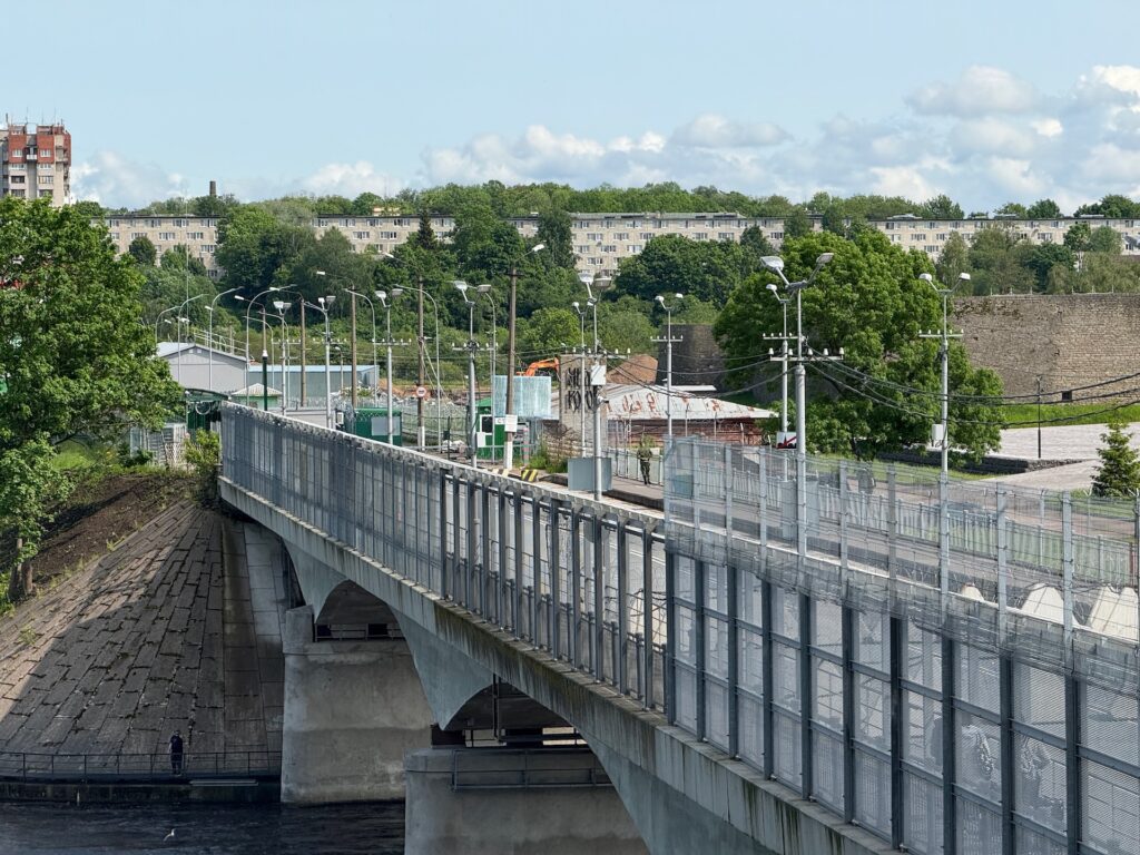Baltikum 2025 – Lettland 2 – 24 Eine Betonbrücke mit Metallzäunen auf beiden Seiten überquert einen Fluss. Im Hintergrund sind Bäume, Wohngebäude und ein teilweise bewölkter Himmel zu sehen.
