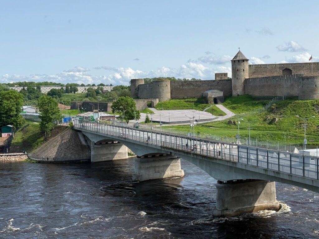 Baltikum 2025 – Lettland 2 – 25 Eine Betonbrücke überquert einen breiten Fluss und führt zu einer historischen Steinfestung mit Türmen und Mauern auf einem grasbewachsenen Hügel unter einem klaren blauen Himmel.