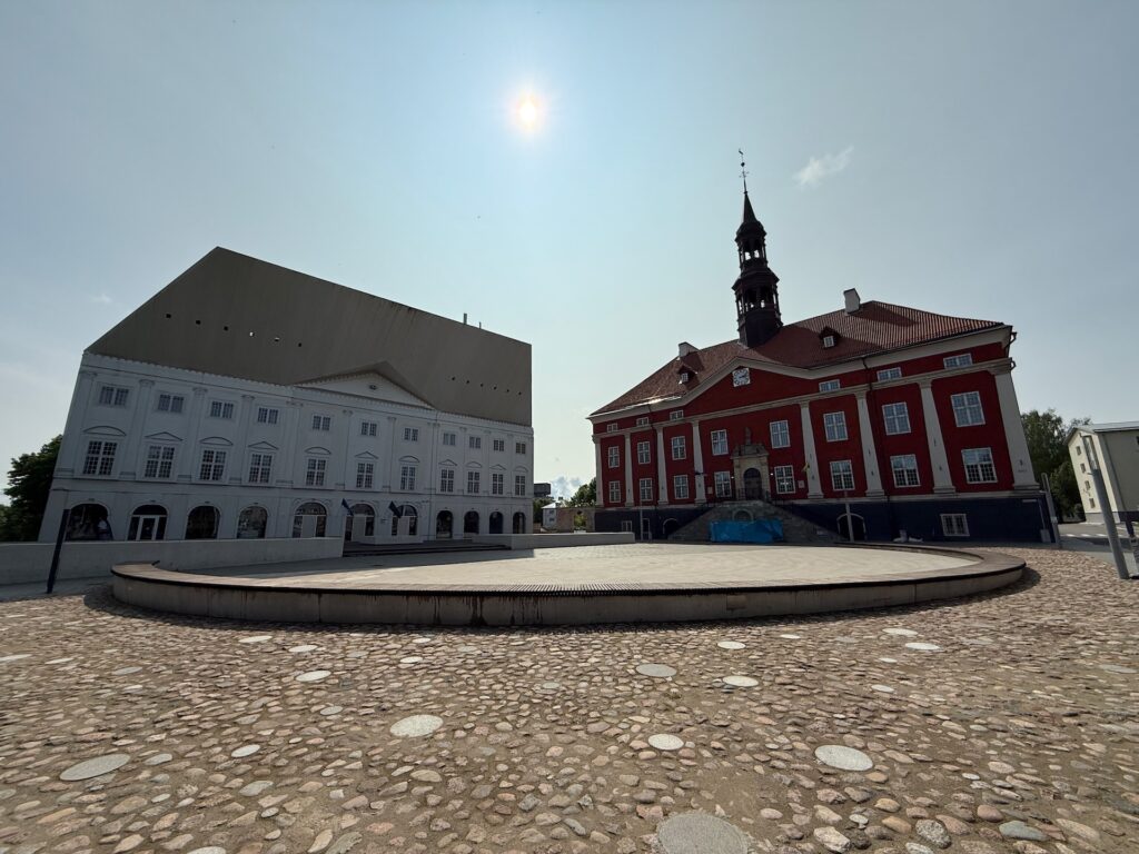 Baltikum 2025 – Lettland 2 – 26 Ein gepflasterter Platz mit einer kreisförmigen Plattform vor zwei Gebäuden: eines ist weiß mit einer modernen oberen Fassade, das andere ist rot mit einem Turm und Bogenfenstern, unter einer hellen Sonne in einem klaren Himmel.