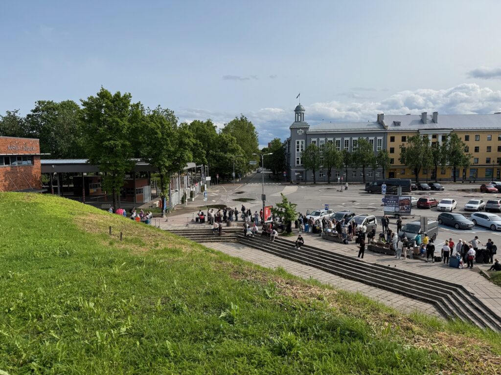Baltikum 2025 – Lettland 2 – 29 Die Menschen stehen auf einer Außentreppe in der Nähe eines grasbewachsenen Hügels Schlange. Im Hintergrund sind unter einem teilweise bewölkten Himmel parkende Autos und ein graues Gebäude mit einem Uhrenturm zu sehen.