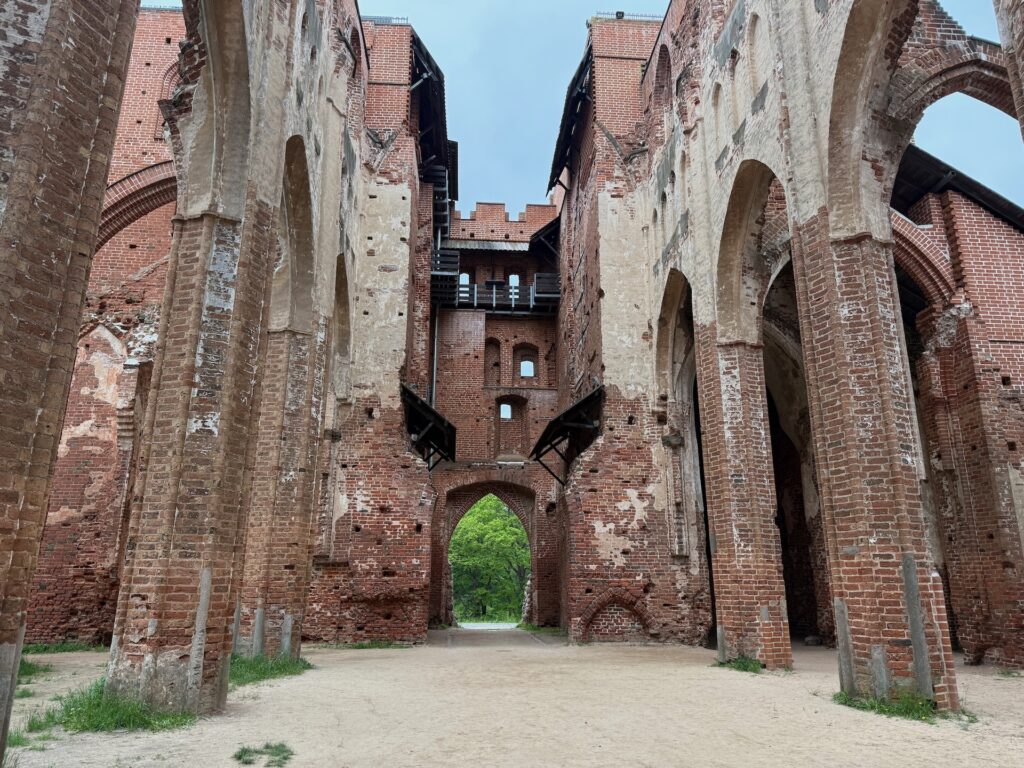 Baltikum 2025 – Lettland 2 – 3 Blick auf ein großes, teilweise zerstörtes Backsteingebäude mit hohen Bögen, verwitterten Wänden und einem offenen Torbogen, der ins Grüne führt. Die Decke fehlt und gibt den Blick auf den Himmel frei.