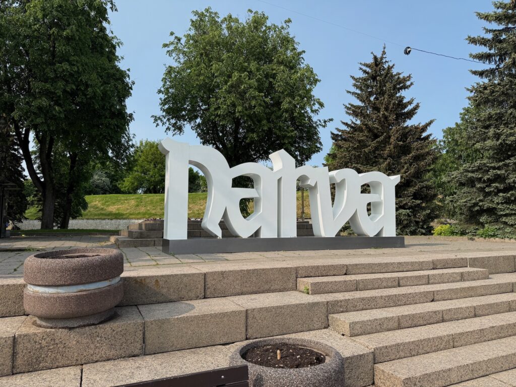 Baltikum 2025 – Lettland 2 – 30 Große weiße Buchstaben mit der Aufschrift „Narva“ stehen auf einer Plattform zwischen Stufen und Pflanzkübeln, umgeben von grünen Bäumen und einer Rasenfläche unter klarem Himmel.