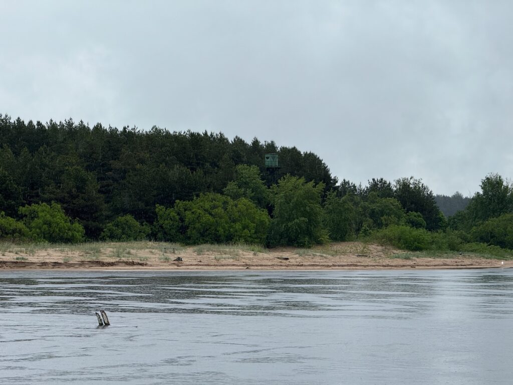 Baltikum 2025 – Lettland 2 – 31 Im Vordergrund fließt ein ruhiger Fluss mit einem sandigen Ufer und dichtem grünen Wald dahinter. Über den Bäumen ist unter einem bewölkten Himmel teilweise ein kleines grünes Bauwerk zu erkennen, möglicherweise ein Aussichtsturm.