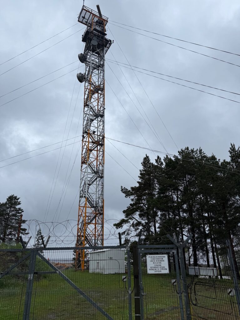 Baltikum 2025 – Lettland 2 – 32 Hinter einem Stacheldrahtzaun und einem Tor steht ein hoher metallener Kommunikationsturm mit Antennen, umgeben von Bäumen unter einem bewölkten Himmel. Am Zauntor ist ein Warnschild angebracht.