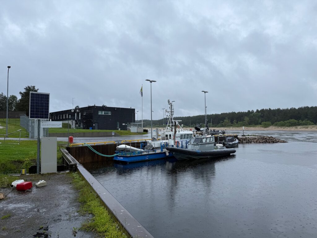 Baltikum 2025 – Lettland 2 – 33 Zwei kleine Boote liegen an einem regnerischen Tag an einem Pier. Im Hintergrund sind ein modernes schwarzes Gebäude, ein Fahnenmast und Bäume unter einem bewölkten Himmel zu sehen. Nasser Boden und einige Solarmodule sind in Ufernähe zu sehen.