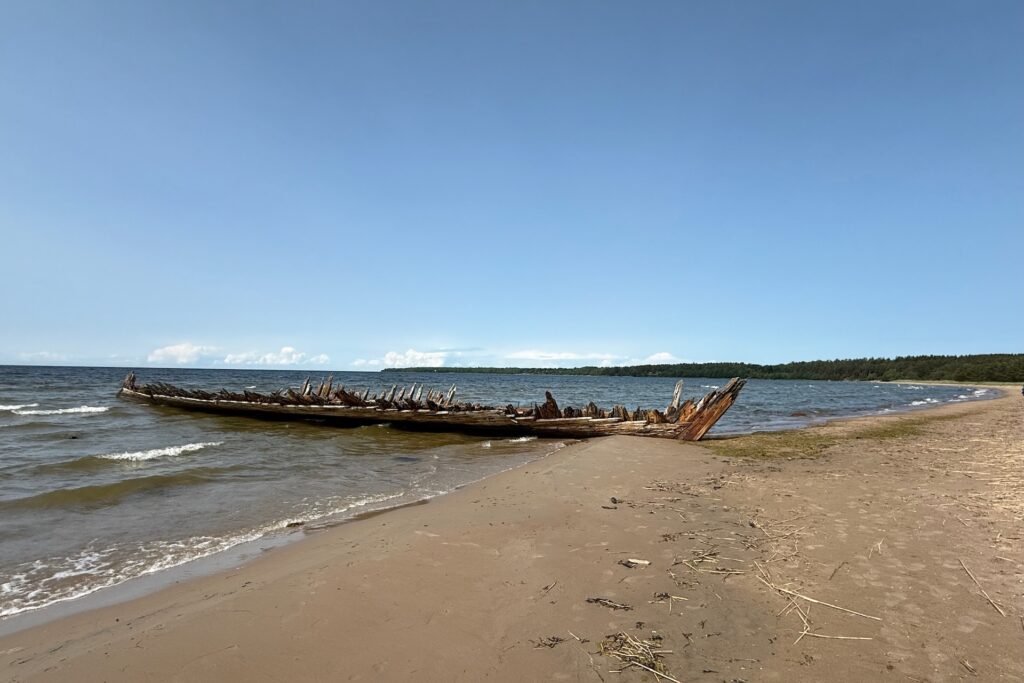 Baltikum 2025 – Lettland 2 – 37 Ein verwittertes Schiffswrack aus Holz liegt teilweise unter Wasser am Rand eines Sandstrandes mit sanften Wellen und einer entfernten, von Bäumen gesäumten Küste unter einem klaren, blauen Himmel.