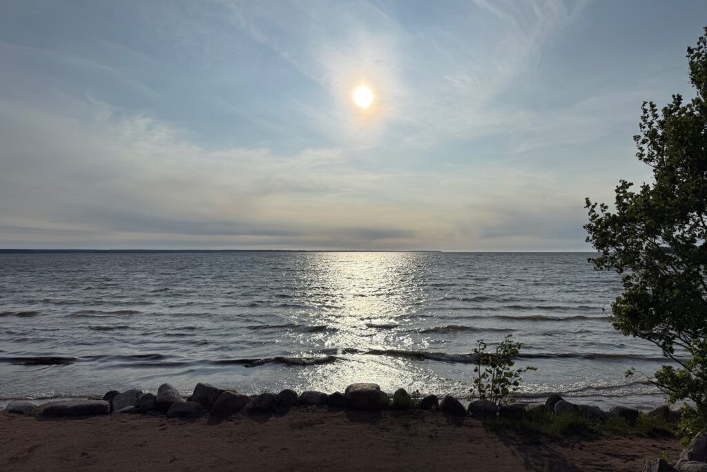 Baltikum 2025 – Lettland 2 – 39 Die Sonne scheint über einen ruhigen See und spiegelt sich auf der Wasseroberfläche. Sanfte Wellen rollen auf ein sandiges, von Felsen gesäumtes Ufer zu. Auf der rechten Bildseite sind einige grüne Bäume zu sehen.