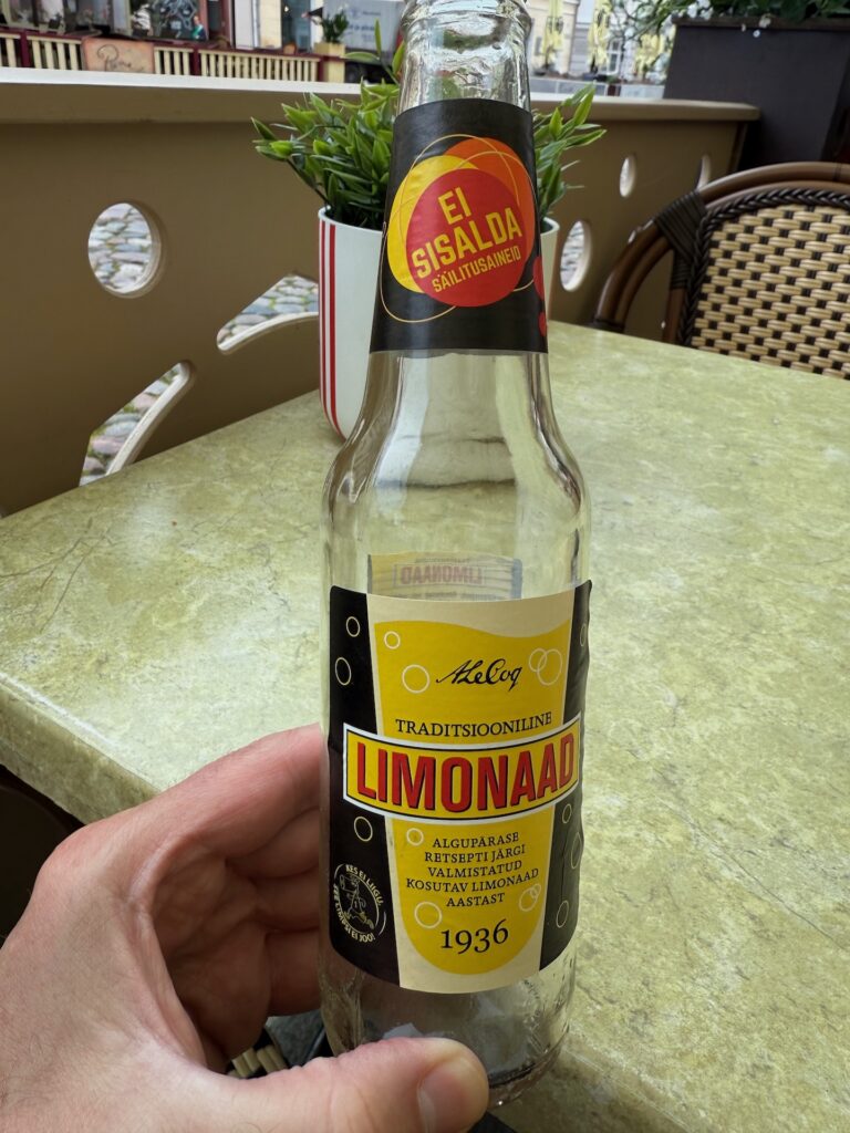 Baltikum 2025 – Lettland 2 – 4 Eine Hand hält eine leere Glasflasche traditioneller estnischer Limonade mit der Aufschrift „LIMONAAD“ in gelbem und schwarzem Design, die auf einem grünen Tisch im Freien in einem Café steht.