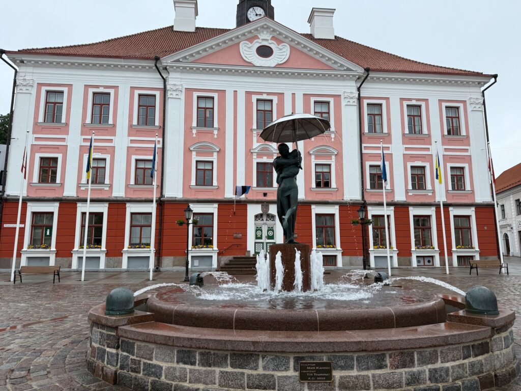 Baltikum 2025 – Lettland 2 – 5 Ein Steinbrunnen mit einer Bronzeskulptur von zwei Menschen unter einem Regenschirm steht vor einem großen, rosa-weißen historischen Gebäude mit einer Uhr an der Spitze. Der Platz ist mit Kopfsteinpflaster gepflastert.