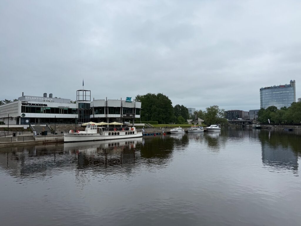Baltikum 2025 – Lettland 2 – 6 Ein Fluss mit Booten, die neben einem modernen Gebäude mit der Aufschrift „Restaurant“ (links) anlegen, im Hintergrund Bäume und rechts ein hohes Gebäude unter einem bewölkten Himmel.