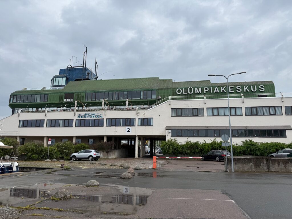 Ein großes, modernistisches Gebäude mit einem grünen oberen Stockwerk und einem weißen unteren Stockwerk, auf dem das Zeichen &quot;OLÜMPIAKESKUS&quot; steht. Der untere Teil ist mit einem &quot;Restoran&quot;-Schild versehen. Das Gebäude steht an einem nassen, bedeckten Tag mit parkenden Autos in der Nähe.