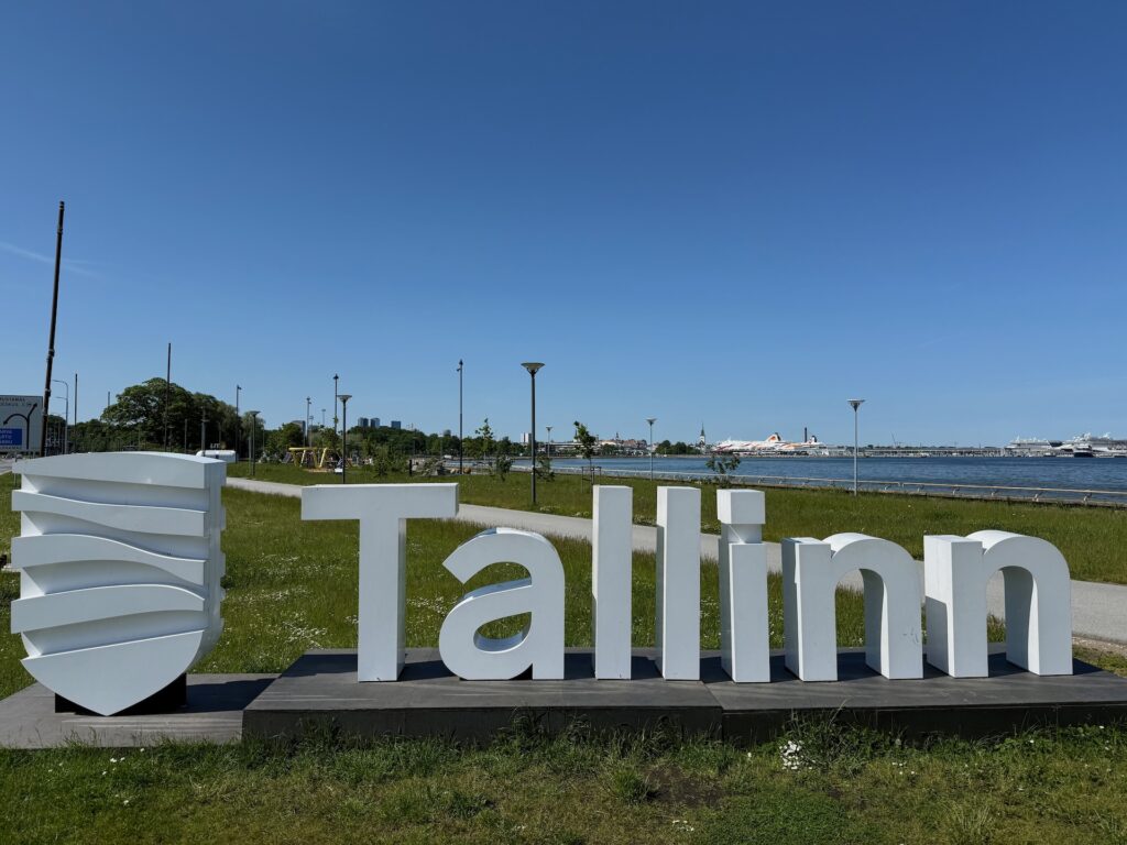 Ein großes weißes &quot;Tallinn&quot;-Schild mit einem Wappen steht auf einer Wiese neben einer Uferpromenade unter einem klaren blauen Himmel. Im Hintergrund sind Bäume, Straßenlaternen und entfernte Schiffe zu sehen.