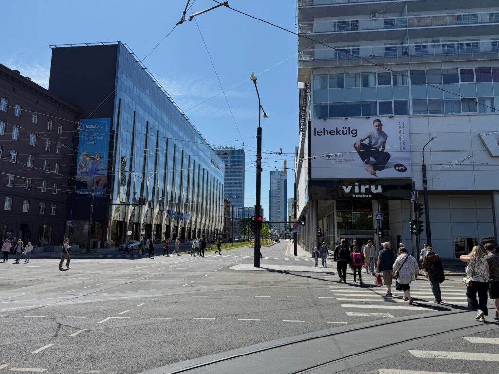 Menschen überqueren an einem sonnigen Tag eine breite Stadtstraße an einem Zebrastreifen, umgeben von modernen und älteren Gebäuden; Straßenbahnschienen und Oberleitungen sind zu sehen, rechts davon ein &quot;viru&quot;-Schild an einem Gebäude.