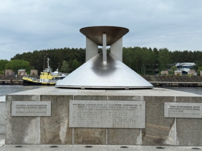 Ein Denkmal aus Beton und Metall mit Tafeln in Form eines Kegels mit einer kreisf&ouml;rmigen &Ouml;ffnung an der Spitze steht an einem Ufer. Im Hintergrund sind ein gelbes Boot und B&auml;ume unter einem bew&ouml;lkten Himmel zu sehen.