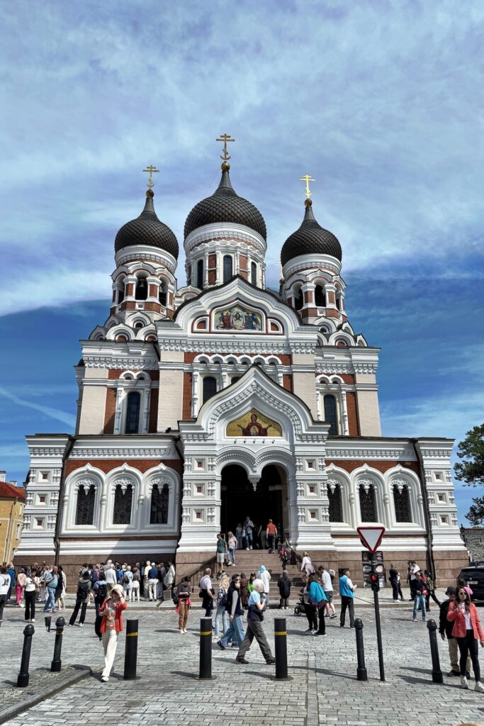 Eine Menschenmenge versammelt sich vor der verschnörkelten Alexander-Newski-Kathedrale in Tallinn, Estland, mit ihren zwiebelförmigen Kuppeln, gewölbten Fenstern und farbenfrohen religiösen Mosaiken vor einem teilweise bewölkten blauen Himmel.