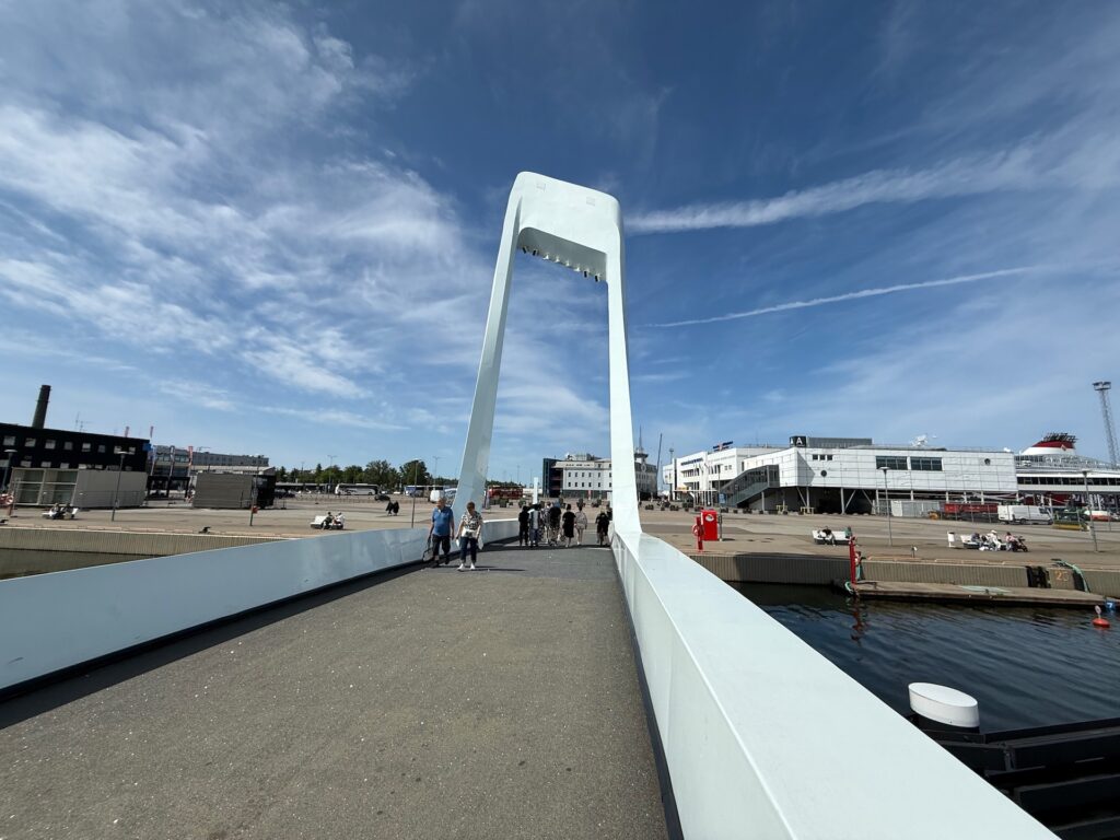 Eine moderne weiße Brücke mit einem Überkopfbogen überspannt einen Kanal und führt zu einem offenen städtischen Bereich, in dem Menschen spazieren gehen und sitzen. Auf der rechten Seite sind moderne Gebäude unter einem blauen Himmel mit Wolkenfetzen zu sehen.