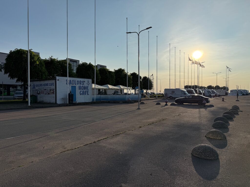 Eine fast leere Straße bei Sonnenuntergang mit Fahnenmasten, einem Gebäude mit der Aufschrift &quot;Sailors Home Cafe&quot;, geparkten Autos, einigen Bäumen und ein paar Wohnmobilen. Die Sonne steht tief am Himmel und wirft lange Schatten.
