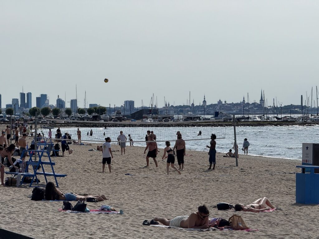 Menschen entspannen sich an einem Sandstrand; einige nehmen ein Sonnenbad auf Handtüchern, während andere Volleyball spielen. Das Meer, Boote und die Skyline der Stadt sind im Hintergrund unter einem leicht dunstigen Himmel zu sehen.