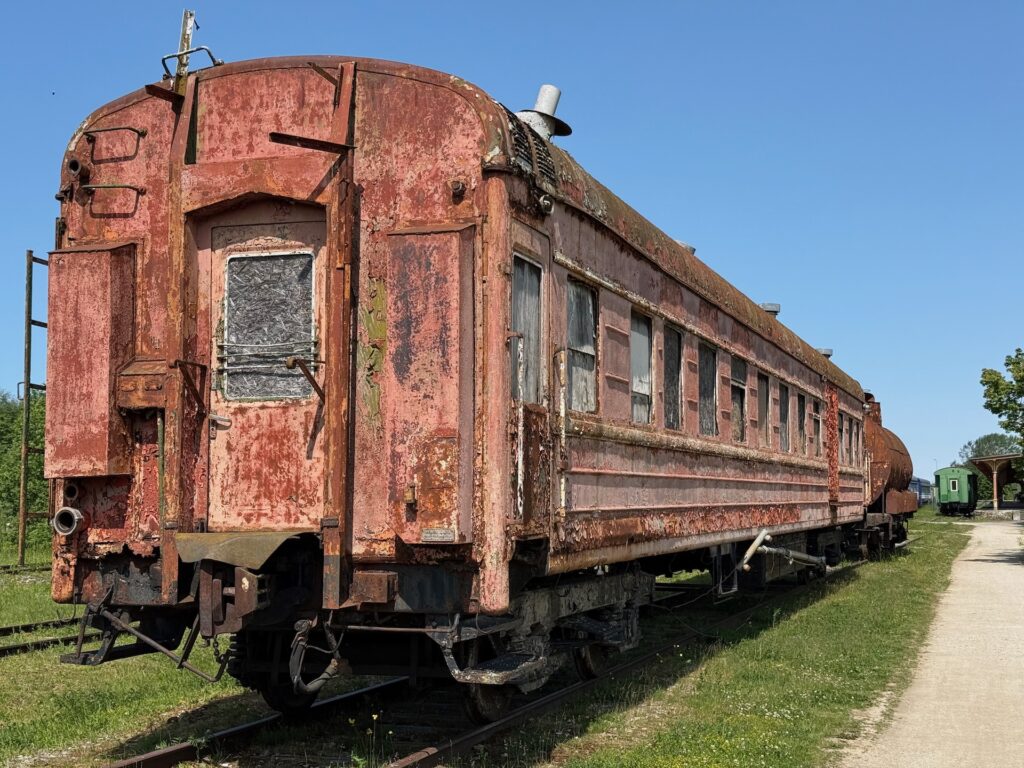 Ein verrosteter, verlassener Zugwaggon steht auf alten Eisenbahnschienen unter einem klaren blauen Himmel. In der Nähe wächst Gras und im Hintergrund ist ein weiterer Waggon zu sehen.