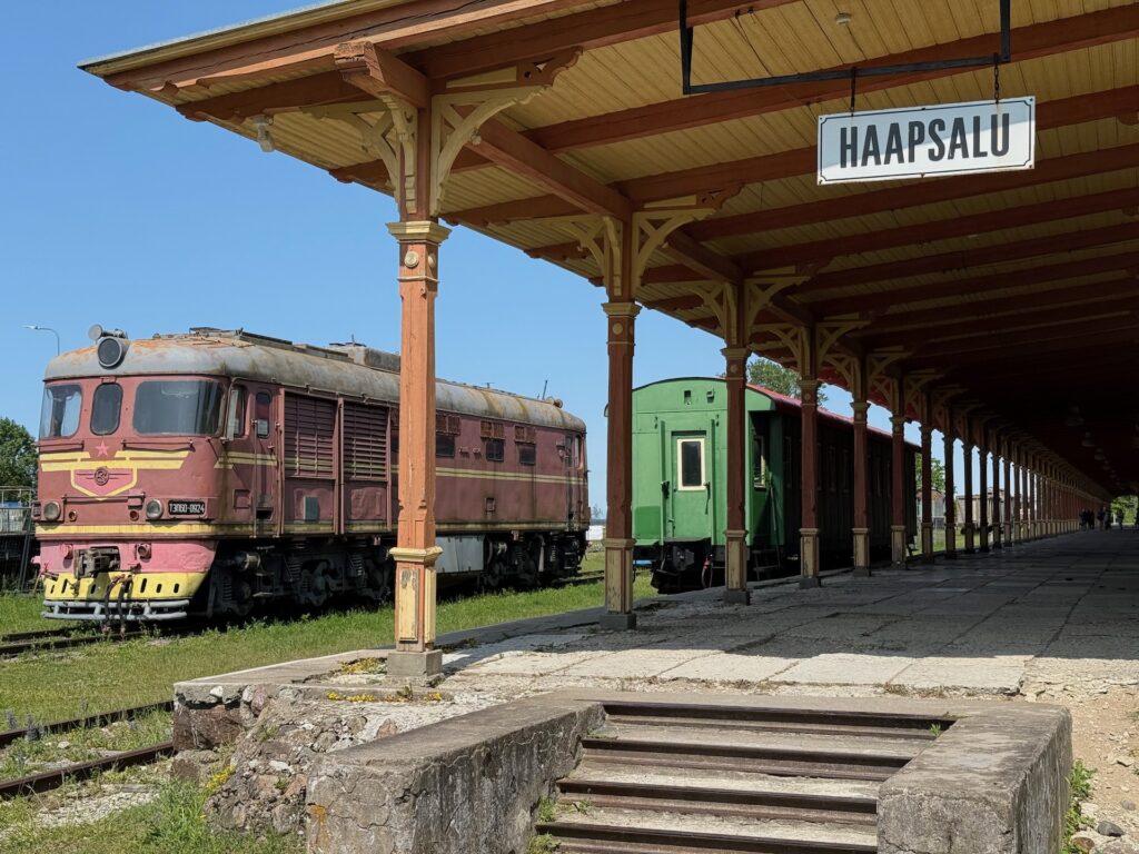 Eine alte rot-gelbe Lokomotive und ein grüner Waggon stehen auf den Gleisen neben einem hölzernen Bahnsteig des Bahnhofs von Haapsalu unter einem strahlend blauen Himmel.