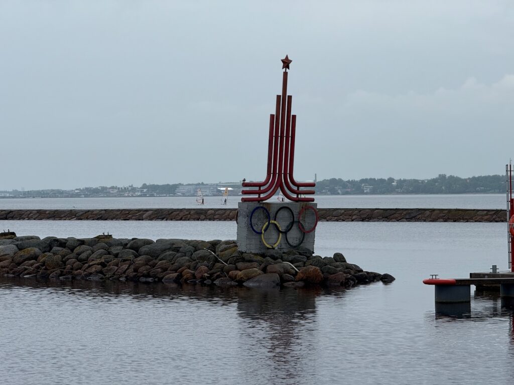 Ein Denkmal mit den olympischen Ringen und einem roten Stern steht auf einem Felsvorsprung im Wasser in der Nähe eines Wellenbrechers, mit einem bewölkten Himmel und einer entfernten Küstenlinie im Hintergrund.