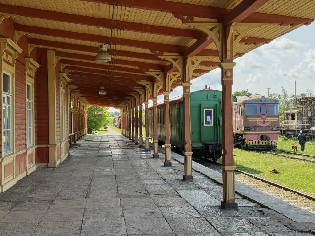 Ein überdachter Bahnsteig mit Holzpfeilern, ein alter grüner Waggon und eine rosa und violette Lokomotive auf parallelen Gleisen unter einem sonnigen Himmel. Im Hintergrund sind Gras und Bäume zu sehen.