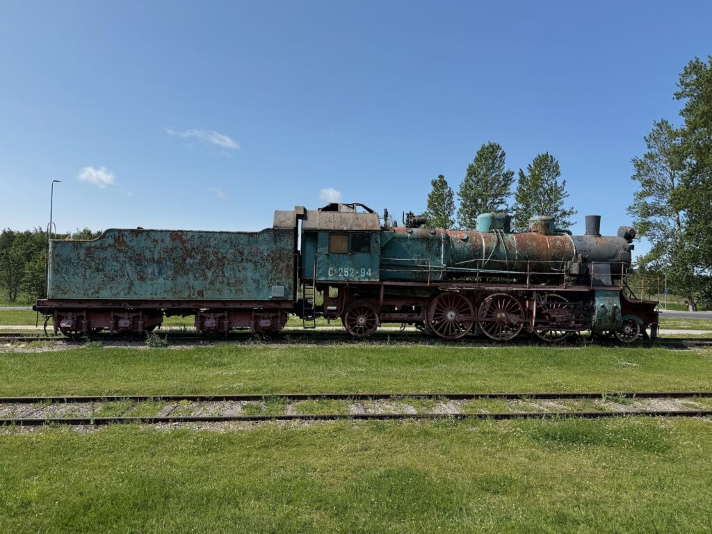 Eine verrostete, alte Dampflokomotive steht auf den Gleisen, umgeben von Gras, mit Bäumen und einem klaren blauen Himmel im Hintergrund.