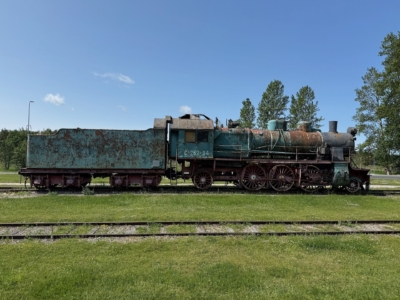 Eine verrostete, alte Dampflokomotive steht auf den Gleisen, umgeben von Gras, mit B&auml;umen und einem klaren blauen Himmel im Hintergrund.