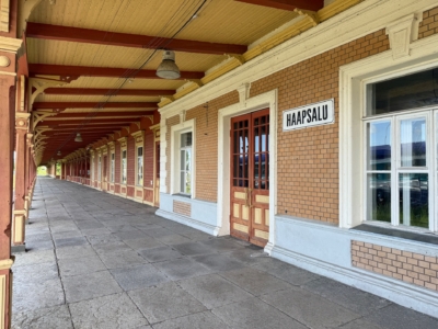 Ein &uuml;berdachter Weg mit Backsteinmauern, Holzpfeilern und einem Schild mit der Aufschrift "Haapsalu" &uuml;ber Doppelt&uuml;ren, wahrscheinlich an einem Bahnhof. Der Bereich ist leer und erstreckt sich in die Ferne.
