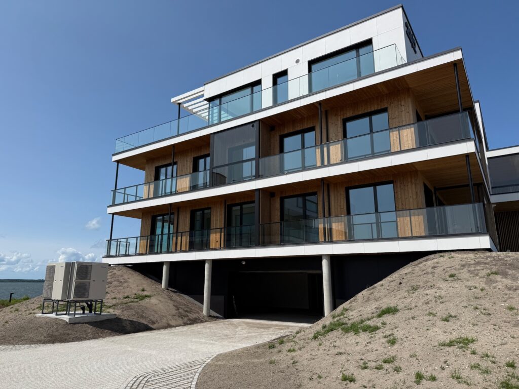 Ein modernes dreistöckiges Wohnhaus mit großen Glasbalkonen, Holzverkleidung und einem ebenerdigen Parkplatz auf einem sandigen Grundstück mit Grasflächen und einem klaren blauen Himmel.