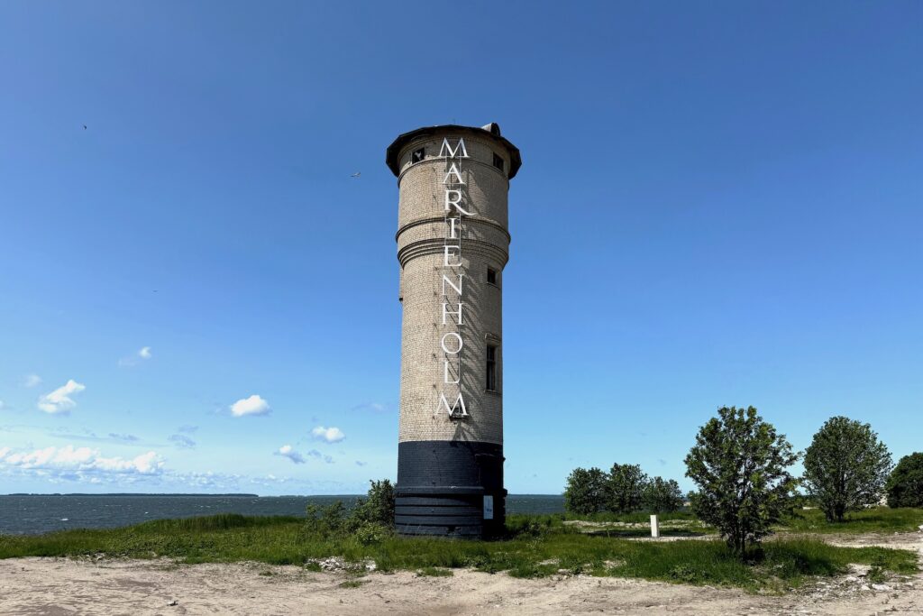 Ein hoher Leuchtturm mit der Aufschrift &quot;MARIENHOLM&quot; steht am Meer, umgeben von Gras, Bäumen und einem blauen Himmel mit vereinzelten Wolken.