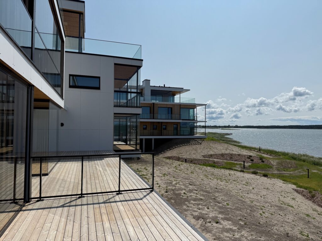 Moderne Wohngebäude mit großen Glasbalkonen überblicken einen Sandstrand und eine Grasfläche neben einem ruhigen Gewässer unter einem klaren, sonnigen Himmel mit vereinzelten Wolken.