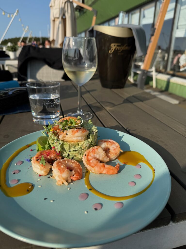 Ein Teller mit Garnelen und Avocadosalat, garniert mit Soßen, steht auf einem Holztisch im Freien. Im Hintergrund stehen im hellen Sonnenlicht ein Glas Weißwein, ein Glas Wasser und ein Freixenet-Weinkühler.