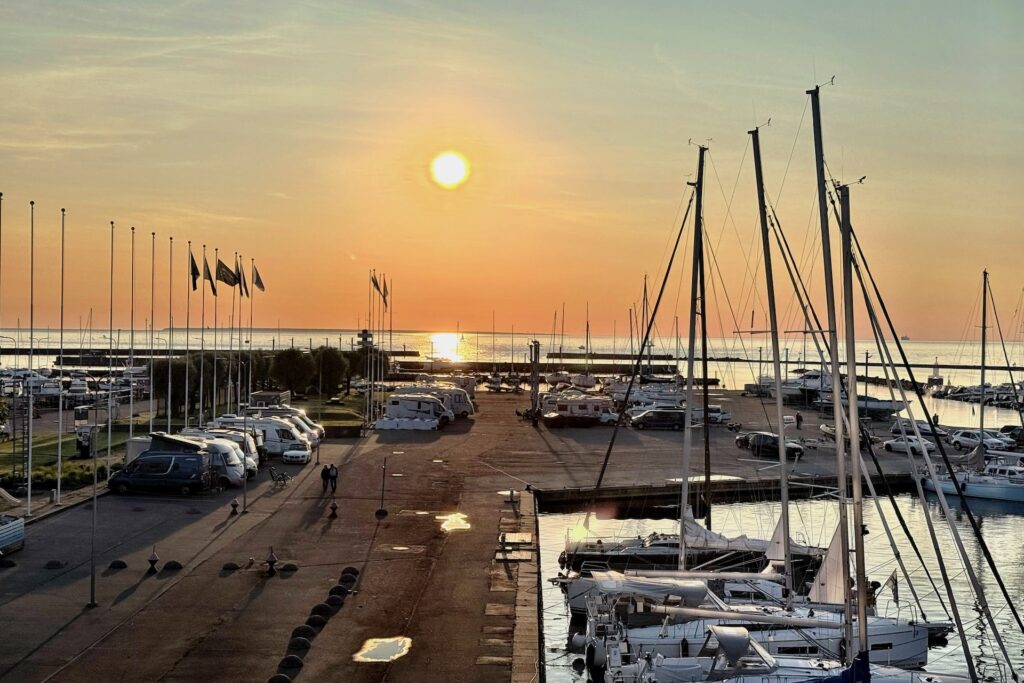 Ein Yachthafen bei Sonnenuntergang mit angedockten Segelbooten, geparkten Autos und Wohnmobilen, Flaggen an Masten und ruhigem Wasser, in dem sich das orangefarbene Licht der Sonne spiegelt.