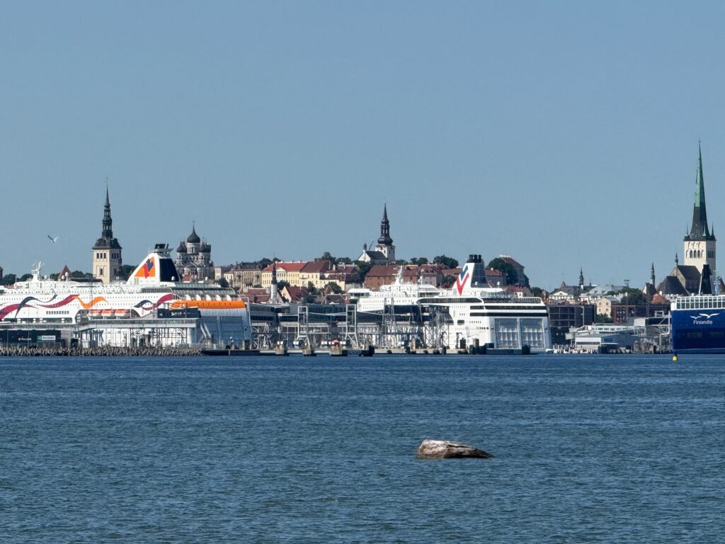 Fähren, die an einem Hafen angedockt haben, mit einer Stadtlandschaft im Hintergrund, einschließlich mehrerer Kirchtürme und historischer Gebäude unter einem klaren blauen Himmel, über ein Gewässer hinweg betrachtet.