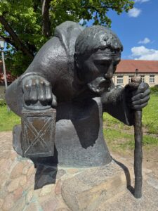 Eine Bronzestatue eines bärtigen Mannes im Umhang, der kniet und in der einen Hand eine Laterne und in der anderen einen Spazierstock hält. Sie steht im Freien auf einem Steinsockel mit Gebäuden und Grün im Hintergrund.