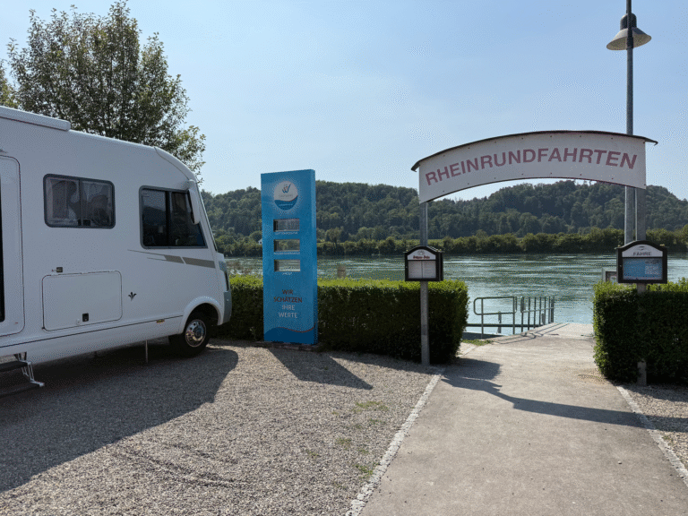 Das Wohnmobil BLUEFIRE - BLUΞFIRΞ parkt neben dem Eingang zu einer Flusstour, auf dem ein Schild mit der Aufschrift "Rheinrundfahrten" steht. Ein blauer Informationskiosk und grüne Hecken säumen den Weg, der zum Ufer führt. Im Hintergrund sind Bäume und Hügel zu sehen.